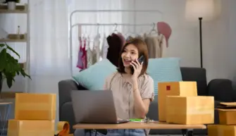Smiling happy young asian woman entrepreneur receiving phone call new sales order boxes product with laptop computer