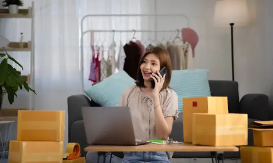 Smiling happy young asian woman entrepreneur receiving phone call new sales order boxes product with laptop computer