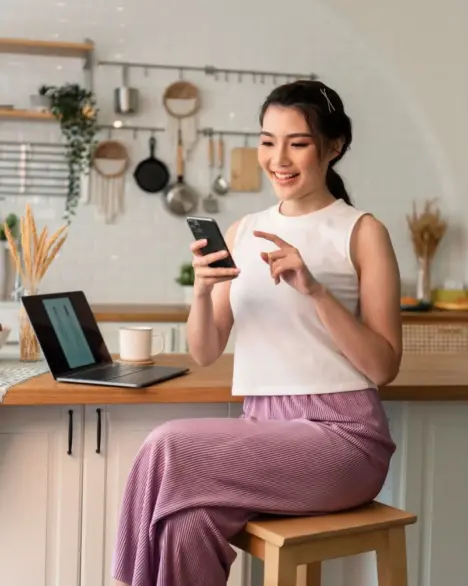 smiling-young-asian-woman-using-mobile-phone-while-sitting-kitchen-room-home-with-laptop Smiling Young Asian Woman Using Mobile Phone While Sitting Kitchen Room Home With Laptop