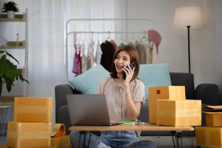 Smiling happy young asian woman entrepreneur receiving phone call new sales order boxes product with laptop computer